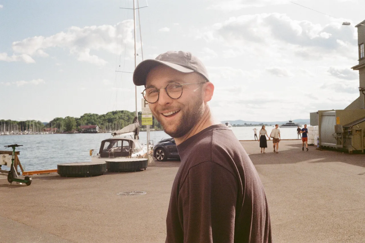 Max Rickards smiling back over his shoulder at a marina, wearing a cap and glasses, with sailboats and a cloudy sky behind him