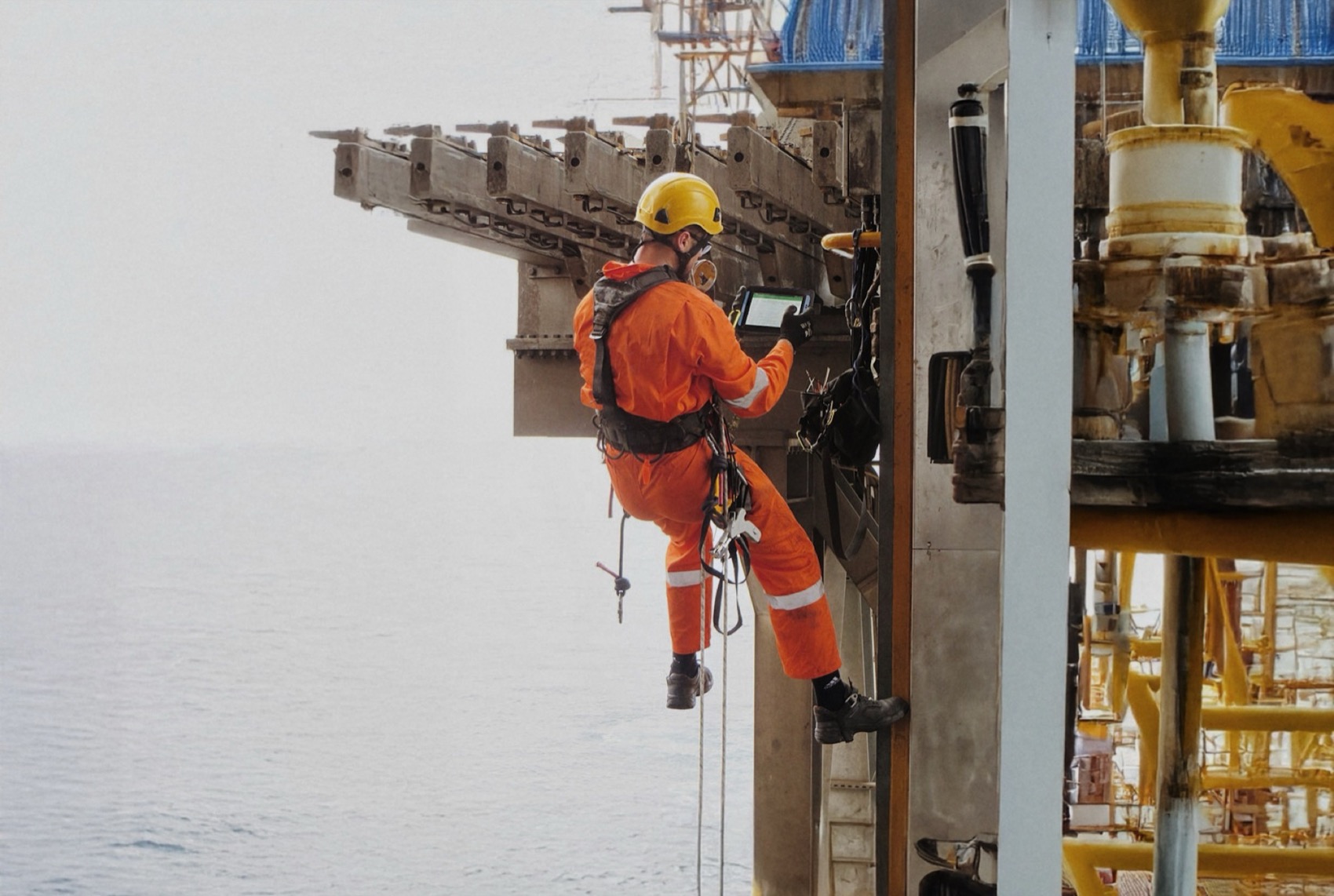 An engineer in a harness and hard hat checking a rugged tablet while working at height on an offshore platform